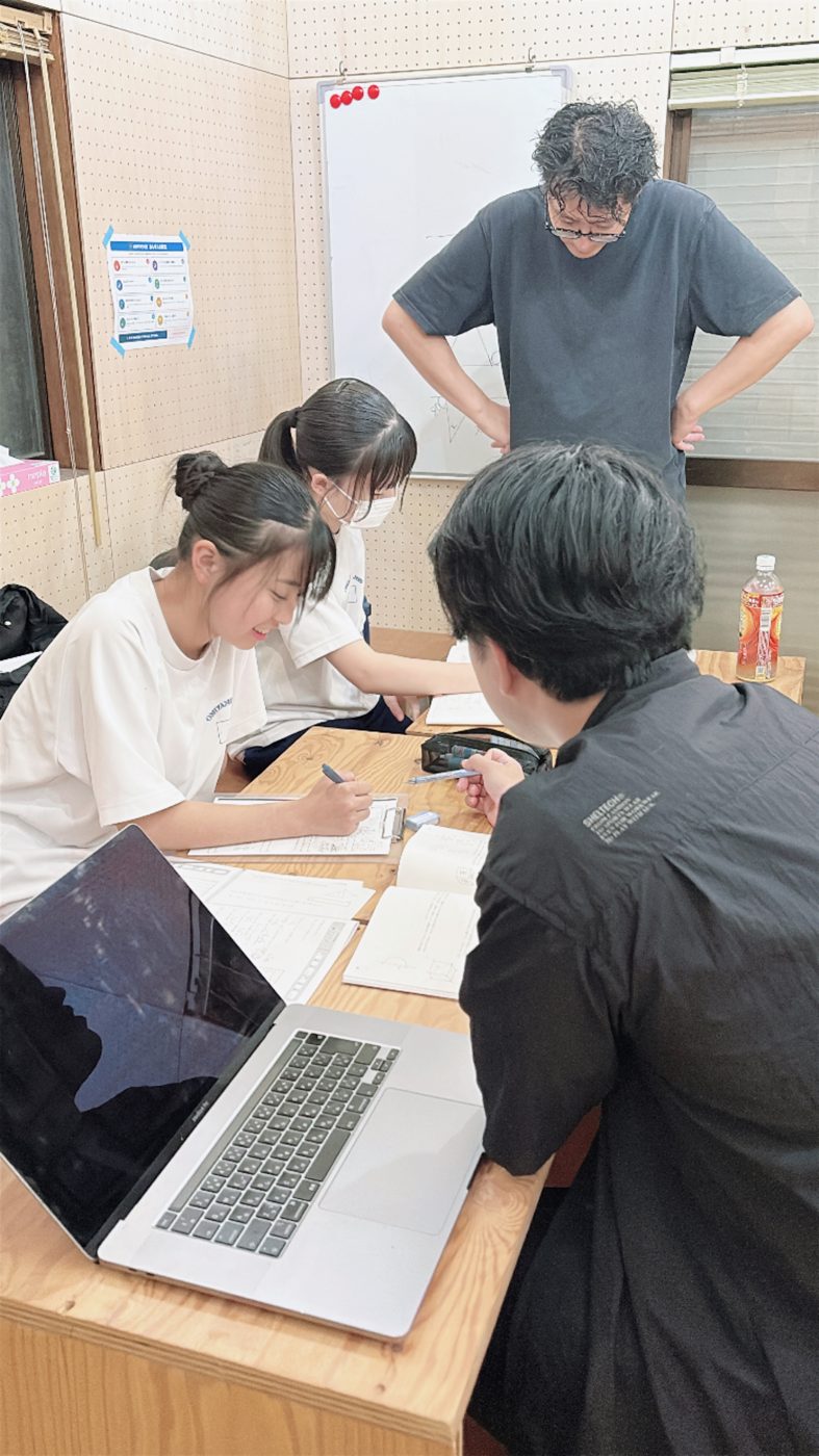 机の横に立った先生と、ノートを見ながら今日やることを決めている生徒